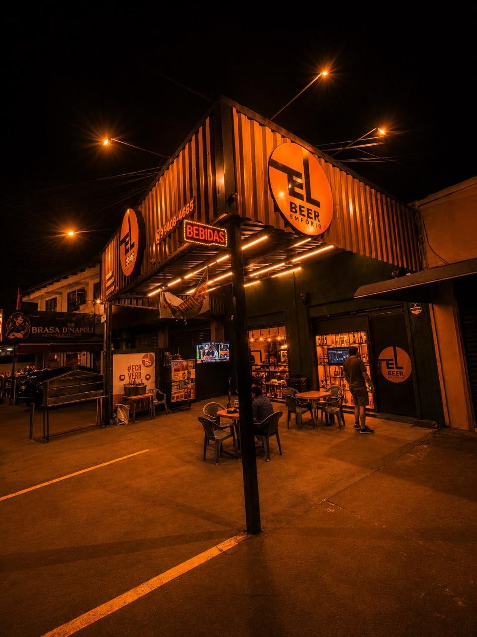 Fachada do El Beer Empório à noite, com o letreiro laranja aceso e mesas na calçada